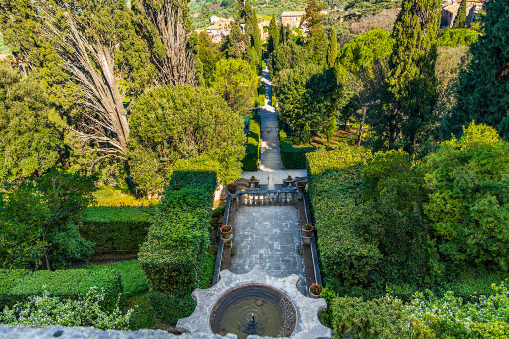 Aerial view of a formal garden with a stone terrace, circular fountain in the foreground, and a long central path flanked by trimmed hedges and tall trees.