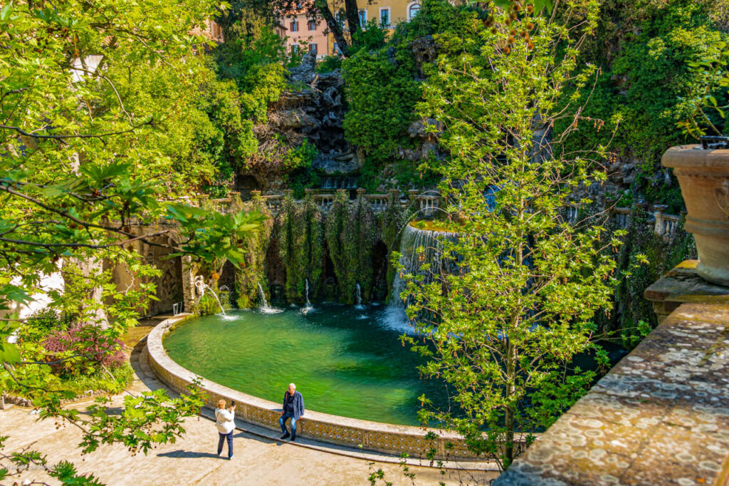 Circular turquoise pool with small waterfalls spilling over ivy-covered walls, framed by trees; two people stand along the railing nearby.