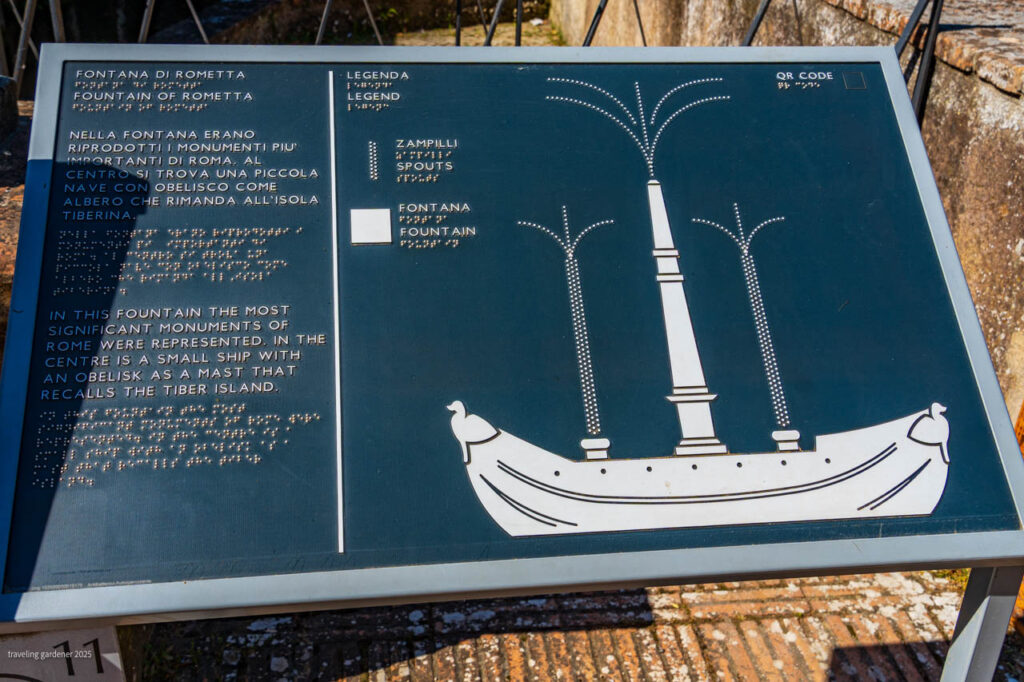 Outdoor information plaque showing a stylized ship fountain diagram with four obelisk-like columns and Italian/English text, mounted on a stand on stone pavement.
