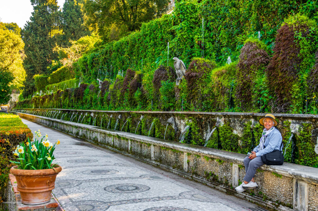 Senior woman in a straw hat sits on a stone bench along a moss-covered fountain wall in a lush formal garden.