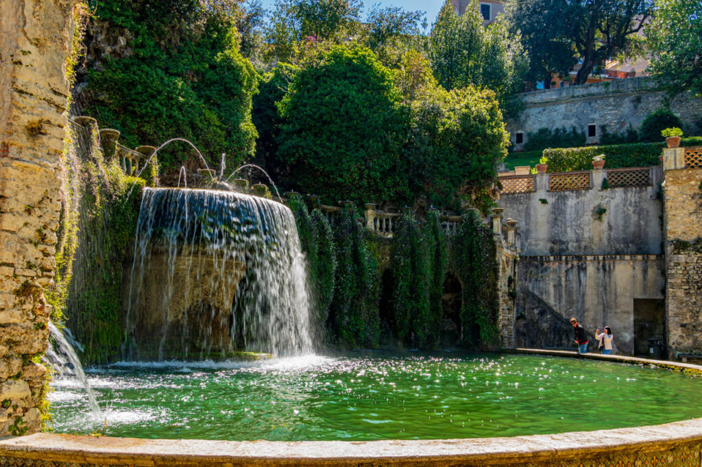 Water cascades from a historic stone fountain into a green circular pool, ivy-covered walls and trees around, with two people near the edge.