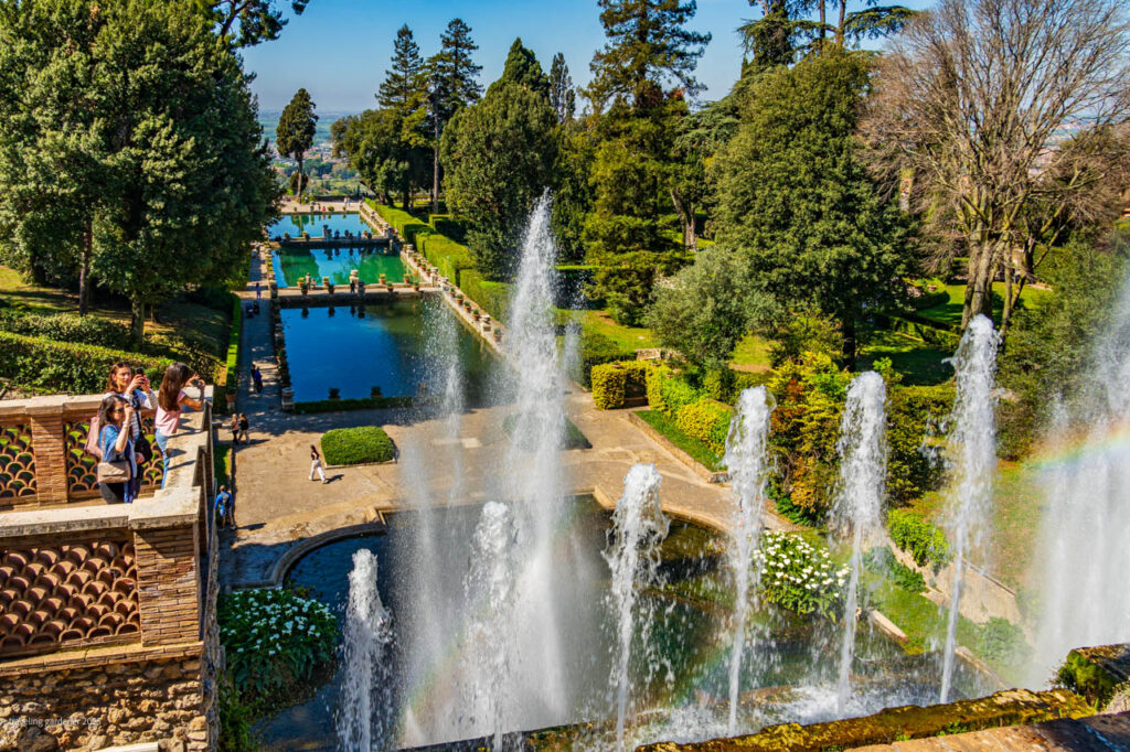 Group of visitors taking photos from a stone terrace overlooking a formal garden with multiple fountains and a long reflecting pool.