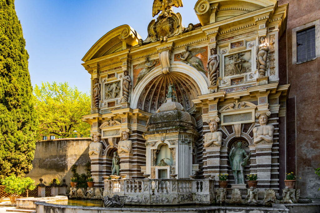 Elaborate historic fountain with arches, columns, and sculpted figures in a sunlit courtyard.
