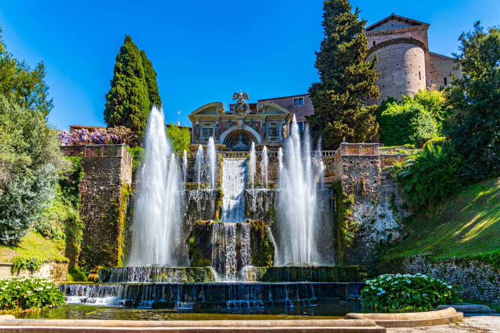 Multi-tiered fountain with cascading water, ornate stone facade, and lush gardens under a bright blue sky.