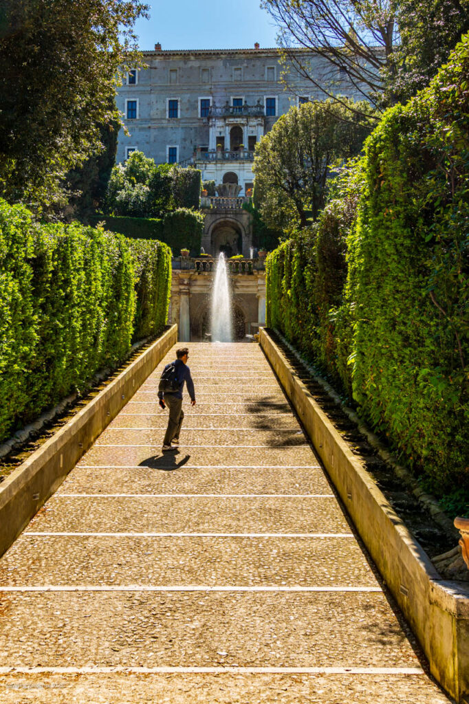 Hiker with a backpack walks up a stone staircase toward a central fountain and a grand historic building framed by tall hedges.