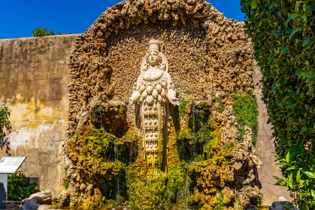 Stone statue of a crowned female figure in a mossy grotto fountain set in a rock wall, with water cascading and a bright blue sky above.