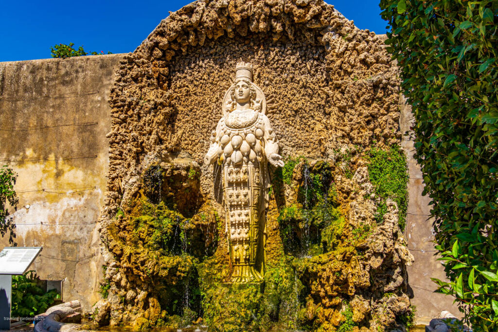 Stone statue of a crowned female figure standing in a mossy fountain carved into a rocky wall with water flowing down the rocks.