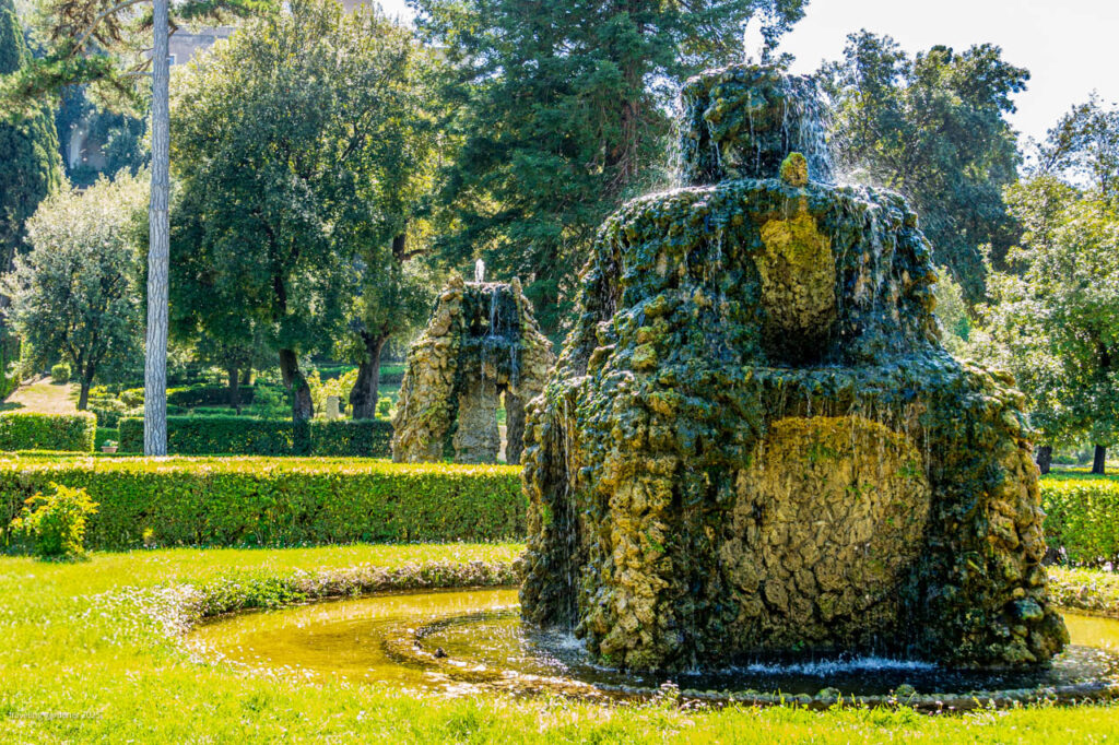 Stone fountain in a formal garden with water trickling down mossy rocks, surrounded by trimmed hedges and trees in a sunny park.
