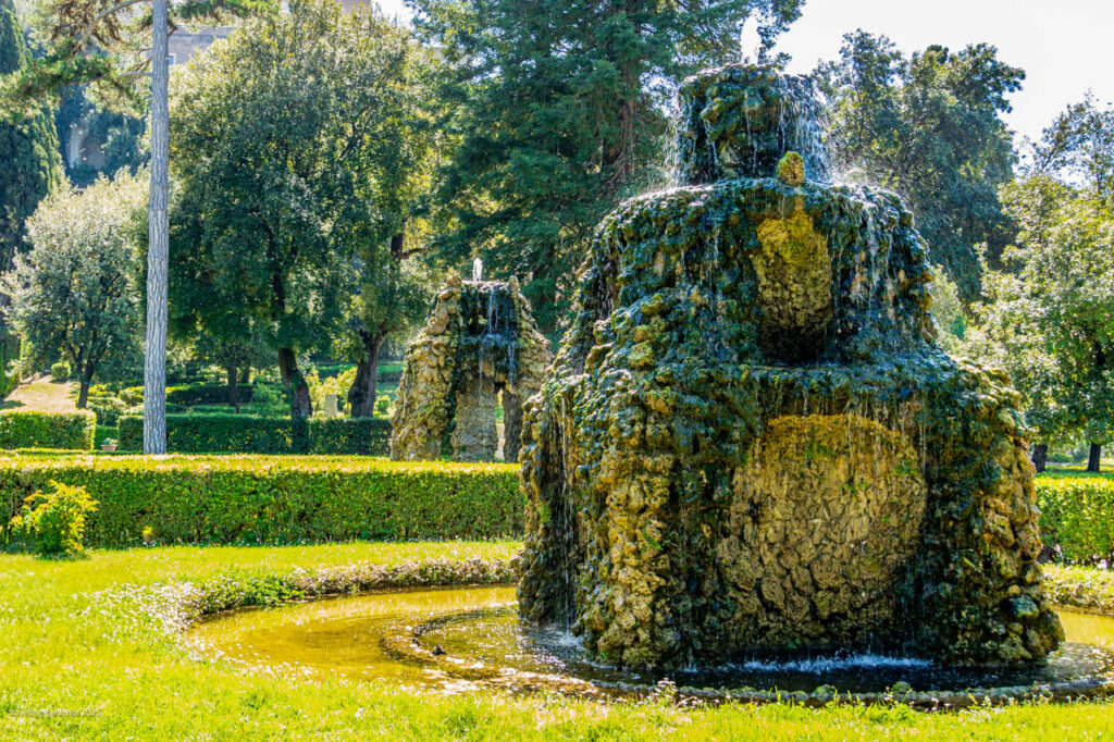 Stone fountain in a manicured garden with water cascading over rocky formations, surrounded by hedges and trees.