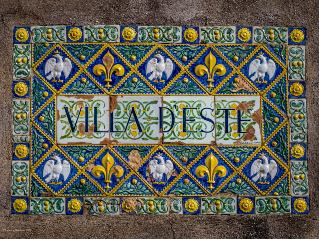 Decorative ceramic tile mosaic in a border frame spelling 'VILLA DESTÉ' with blue diamonds, gold fleur‑de‑lis, and white roosters on blue backgrounds.