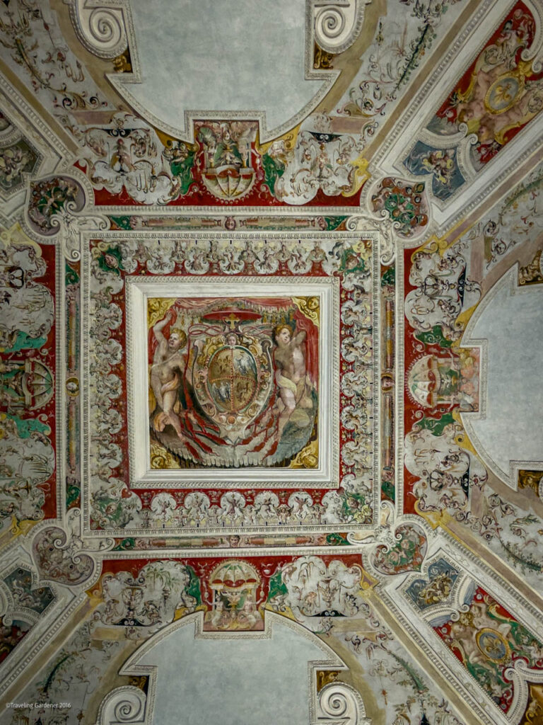 Intricate frescoed ceiling with central square panel featuring classical figures and shields, surrounded by ornate scrollwork and murals on red, green, and gold.