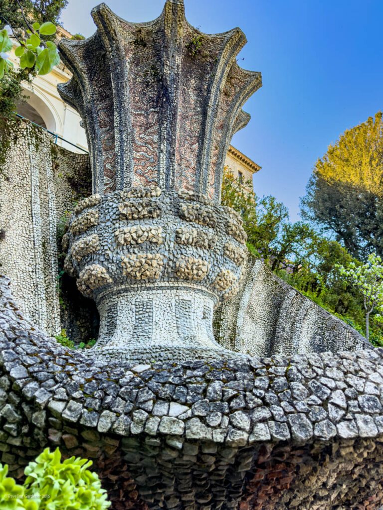 Gaudí-inspired mosaic fountain with petal-like columns rising from a rounded, textured stone base in a garden setting under a blue sky.