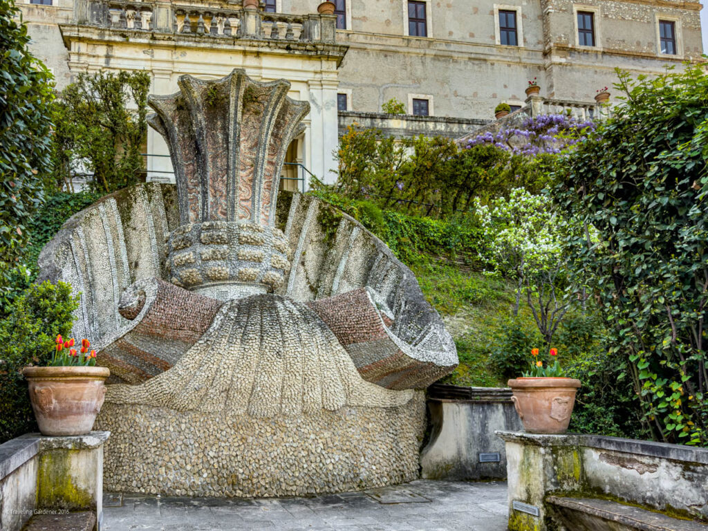 Mosaic stone fountain resembling a blooming flower in a sunlit garden courtyard, with potted tulips and a historic building in the background, ivy-covered walls nearby.