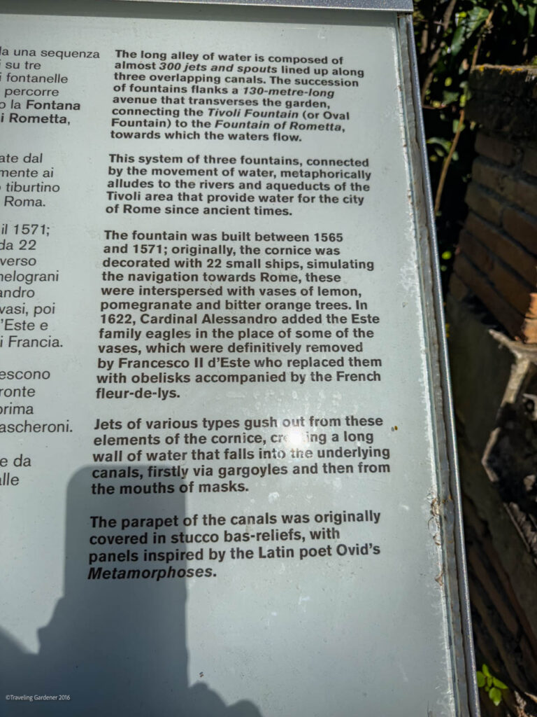 Close-up of a gray metal plaque with dense English text describing a fountain complex and canal system, mounted on a wall.