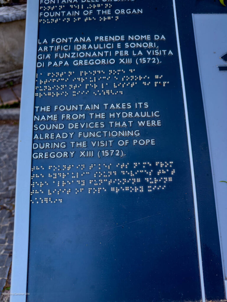 Blue vertical information plaque with raised text in Italian and English and a Braille section, explaining the fountain's name and its link to hydraulic sound devices and Pope Gregory XIII (1572).