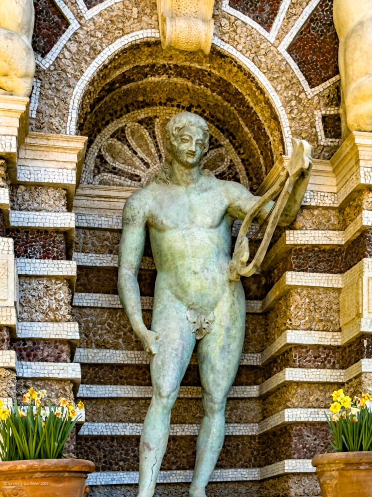 Bronze-green statue of a nude man posing with a draped cloth, set against a mosaic arch backdrop.
