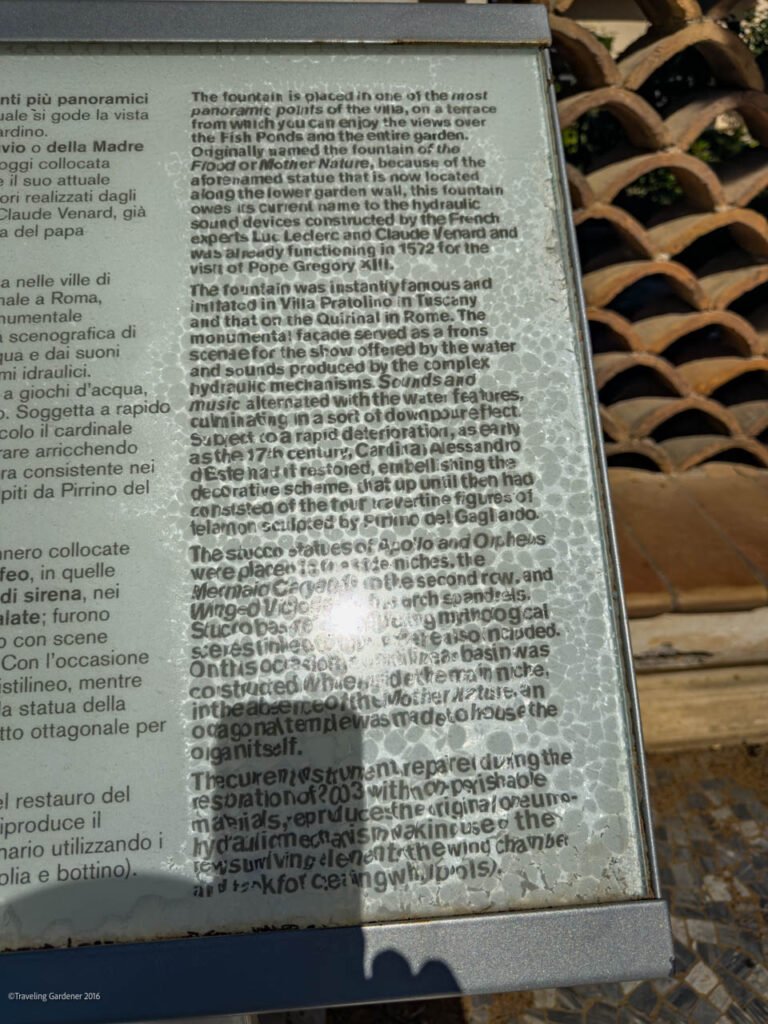 Outdoor metal information plaque with dense multilingual text panels, shadow across the surface, mounted on a blue frame in a garden setting.