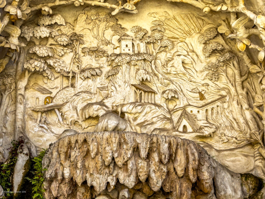 Intricate stone relief showing a hillside village with trees, small houses, a waterwheel and a stream, carved into a cliff with rugged rock textures in the foreground.