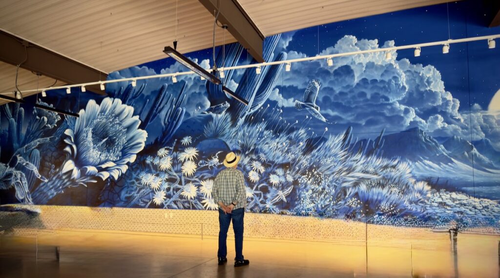 Man wearing a hat stands in front of a large blue mural of flowers inside a gallery or studio. Sky and clouds visible above the mural.