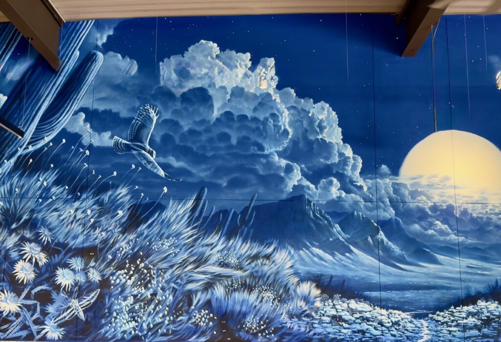 Blue wall mural depicting a dreamy landscape: a setting sun over mountains, billowing clouds, and a foreground of tall grasses and flowers in cool blue tones.