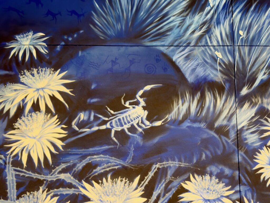 Blue mural with a white scorpion at the center, surrounded by large yellow daisy-like flowers on a blue background.