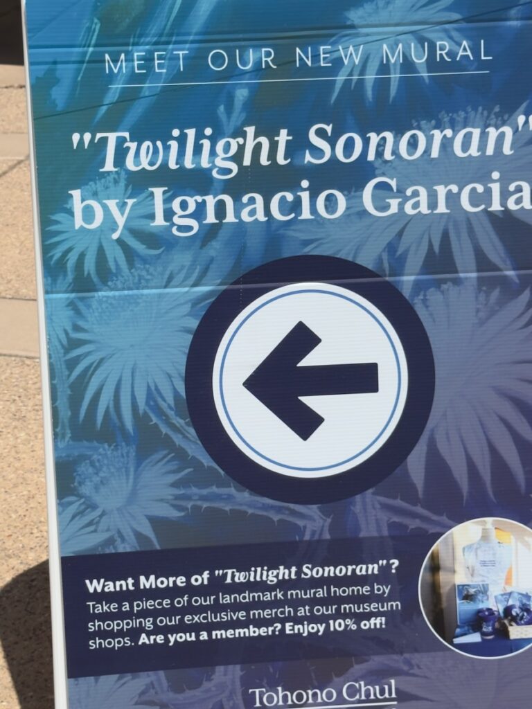 Blue promotional sign advertising the mural 'Twilight Sonoran' by Ignacio Garcia, with a left-pointing circular arrow on a tropical blue background.