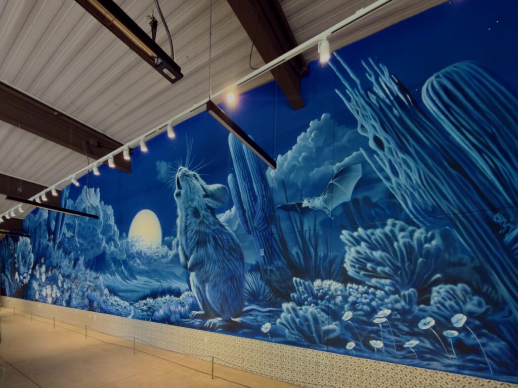 Blue gallery mural of a rabbit‑like creature on a blue coral reef with a rising sun, under ceiling lights.