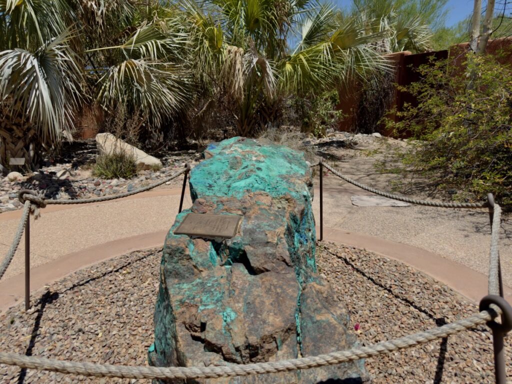 Rock monument with a small plaque surrounded by a rope barrier in a desert garden with palm plants and pebbles.