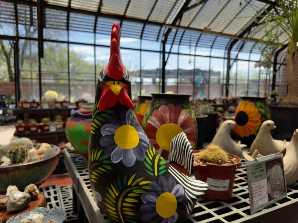 Colorful hand-painted ceramic rooster with blue and yellow flowers on a stand, surrounded by pots in a greenhouse.