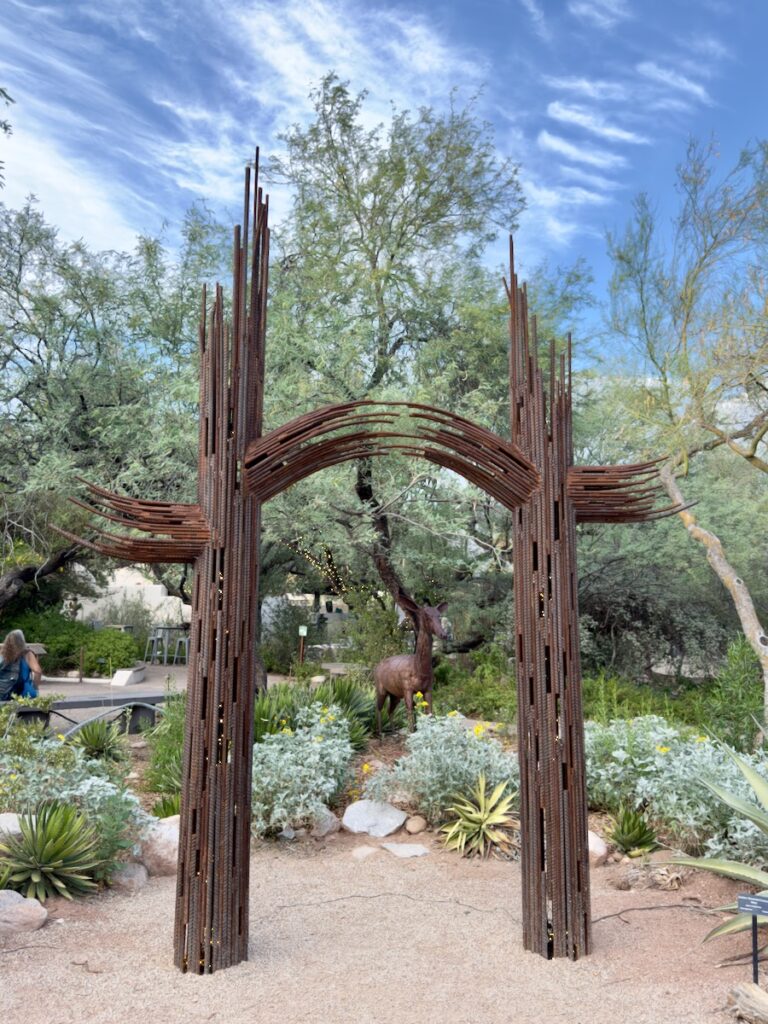 Rusty metal arch gateway sculpture in a desert garden, with a deer statue visible in the background.