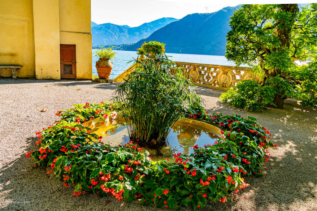 Circular fountain with a tall bamboo plant surrounded by a ring of red flowers, with a scenic lake and mountains in the background.