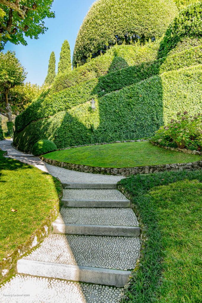 Stone steps wind through a formal hedge garden under a bright blue sky, with trimmed bushes and a pebble path nearby.