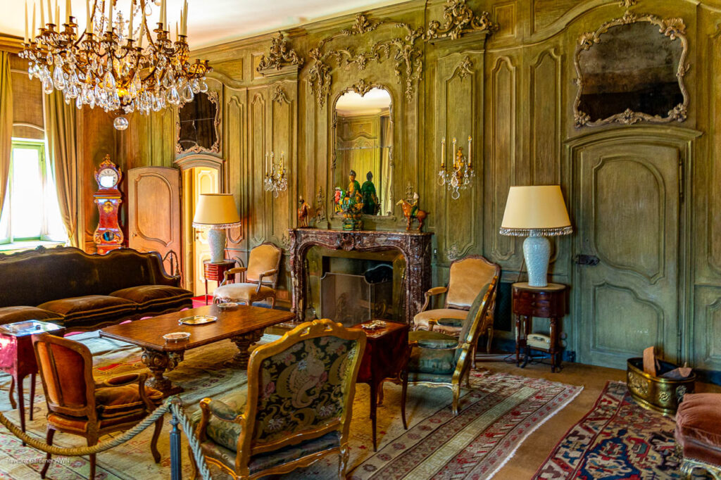 Luxurious antique living room with green paneled walls, crystal chandelier, ornate fireplace mantel, and vintage seating around a wooden table