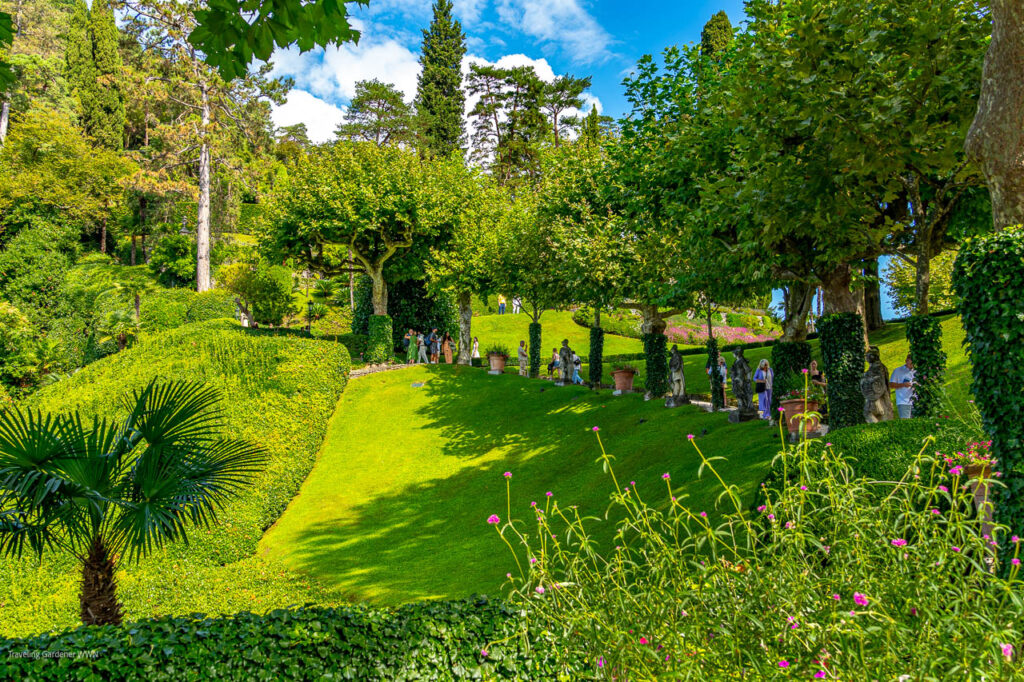 Sunlit formal garden with a grassy slope, trimmed hedges, and a row of statues under trees as visitors stroll along a path.