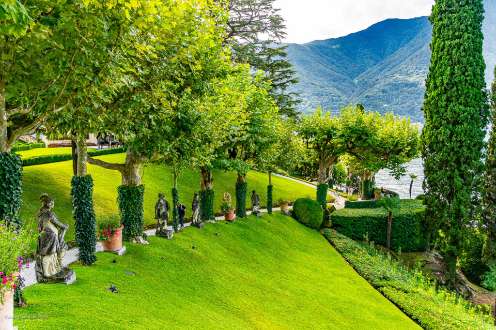 Lush terraced garden with statue sculptures along a trimmed lawn overlooking a lake and mountains in the distance.