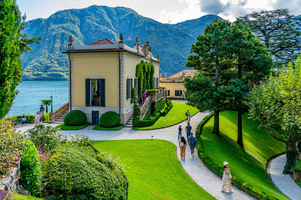 Lakeside villa with yellow walls and green shutters, manicured garden, and winding gravel path with people walking toward the building against a backdrop of mountains and the lake.