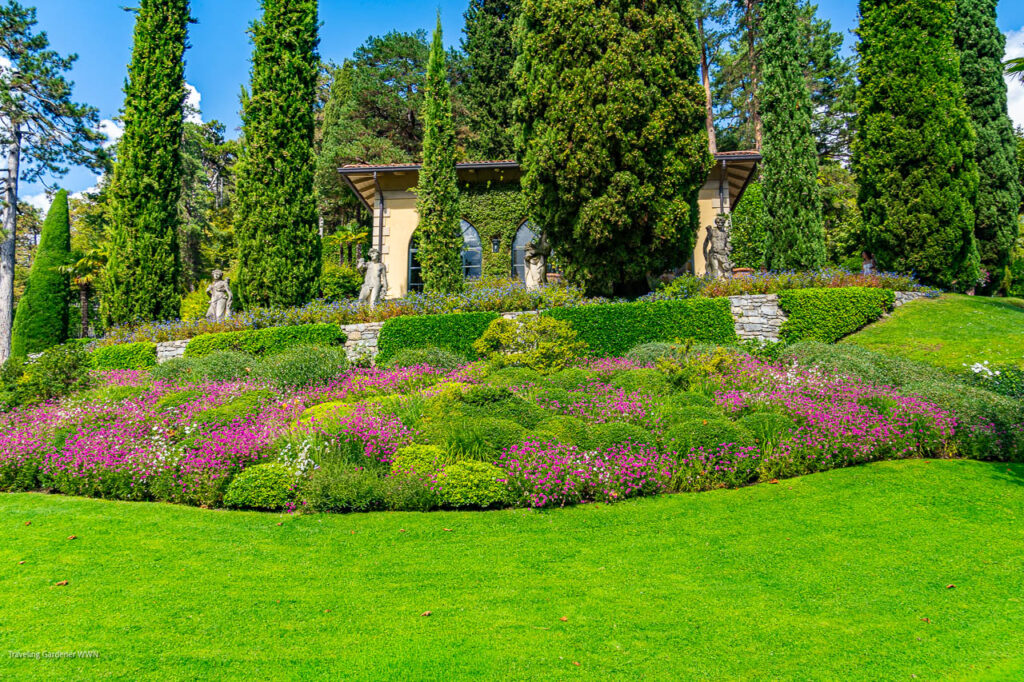 Formal garden with vibrant flower beds, trimmed hedges, statues, and an ivy-covered building in the background.