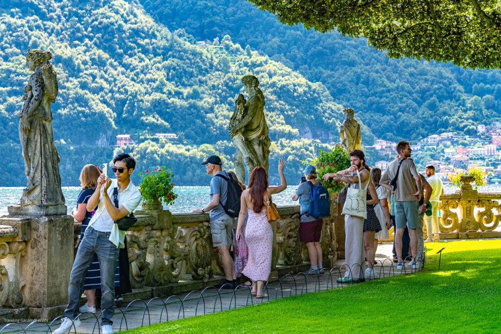 Group of tourists on a stone balustrade by a lake, taking photos with forested hills in the background and statues in the foreground.