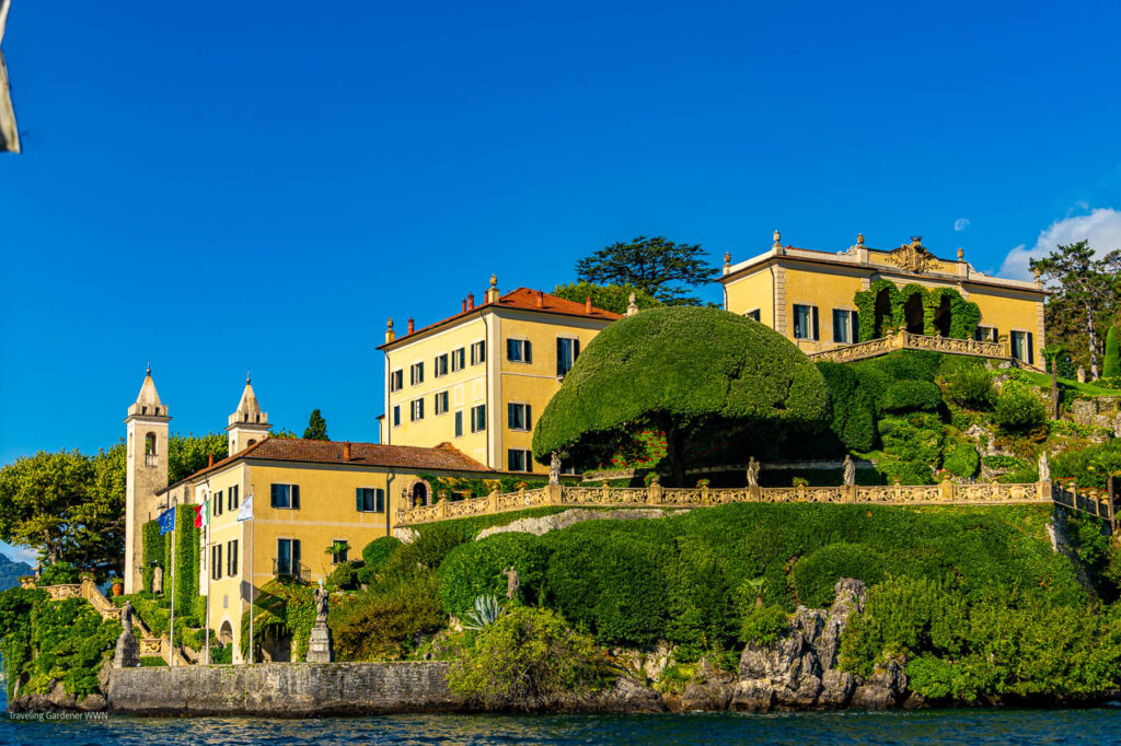 Yellow villa perched on a hillside with manicured topiary and stone balustrades overlooking a lake.