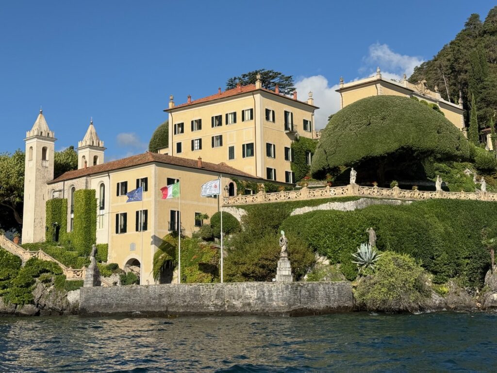 Stately yellow villa by the water with flagpoles, formal gardens, and stone terraces along the lakeshore.