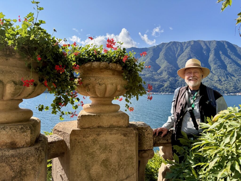 Smiling man in a wide-brimmed hat and vest stands at a stone balustrade overlooking a blue lake and mountains, with red flowers in planters above him