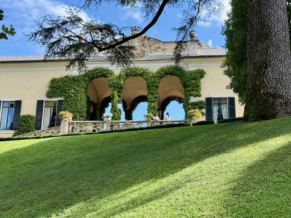 Cream-colored historic terrace with ivy-covered arches and decorative railing; people stand under the arches on a sunny day, with a green lawn in the foreground.