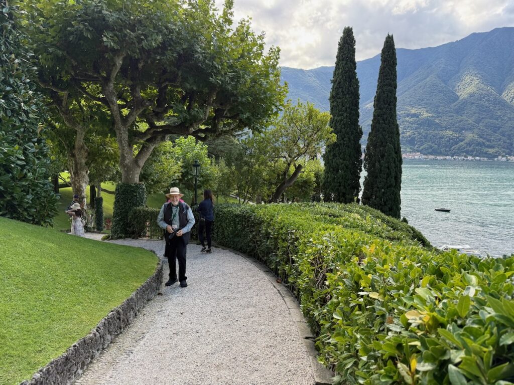 Gravel garden path by a lake with lush hedges and tall trees; a man in a hat walks toward the camera, mountains in the distance.